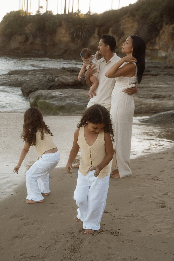 A family of five in cream and butter yellow linen walks the sand at Shaw's Cove in Laguna Beach, the two older girls leading the way
