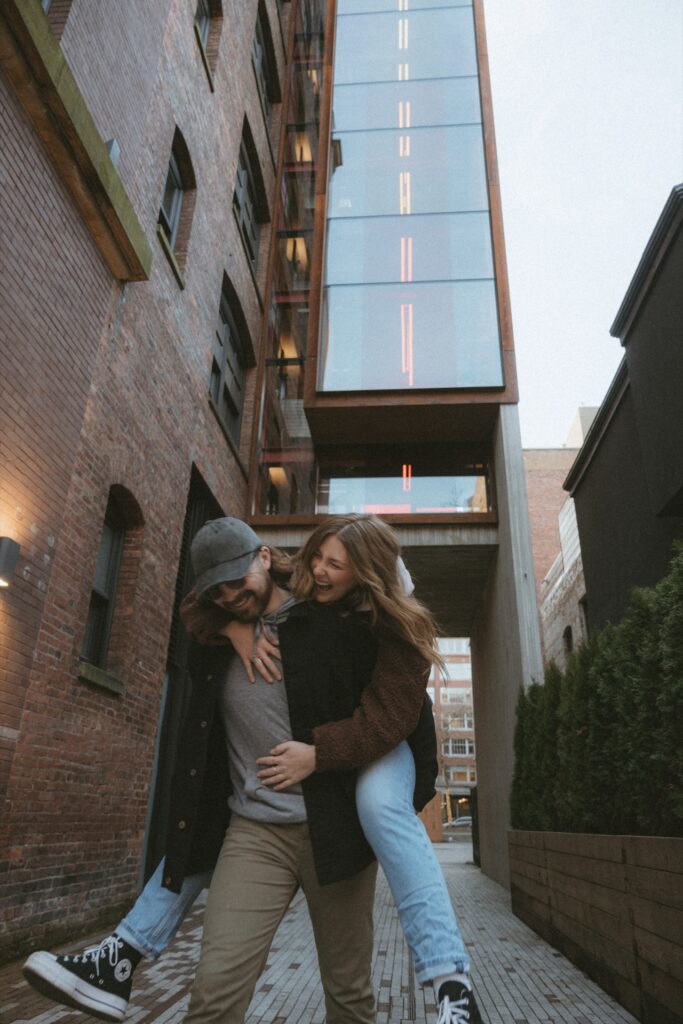 A portrait of a male and a female couple doing a piggyback ride against  brick building near Pioneer Square, Seattle.