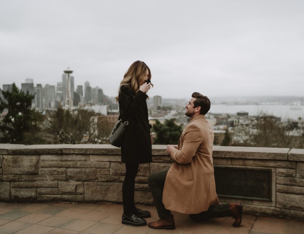 A couples portrait of a male wearing brown coat kneeling at Kerry Park proposing to female near Seattle, WA
