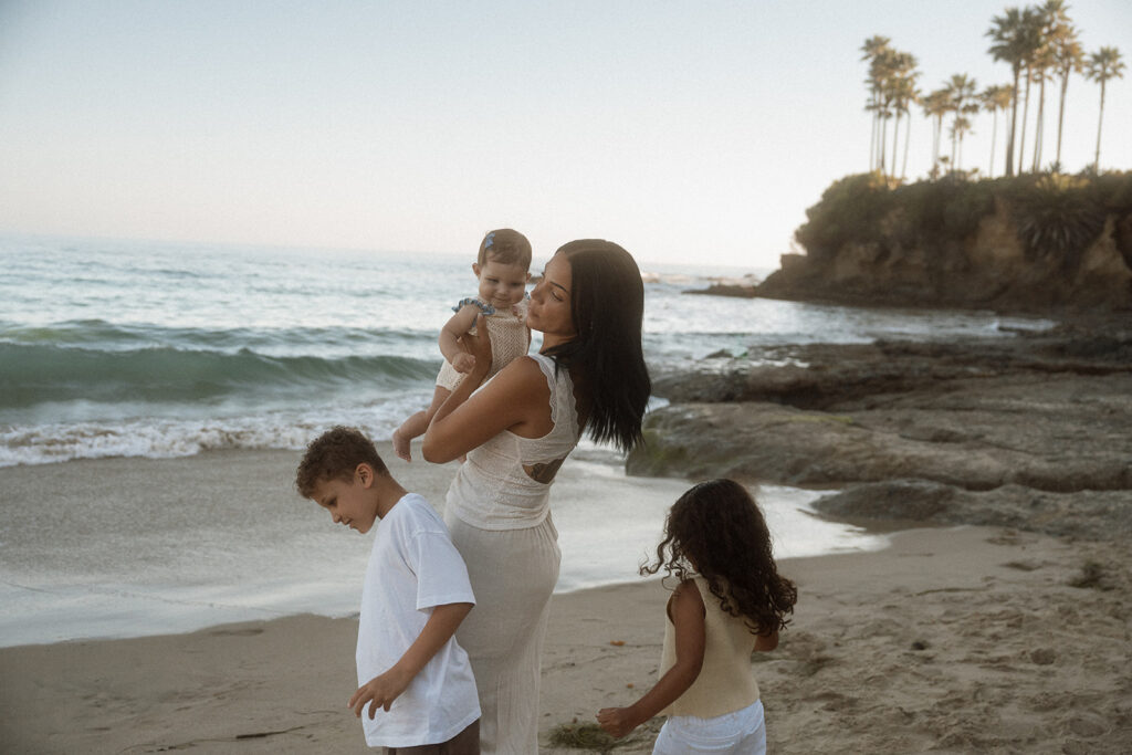 A mother in a cream linen dress carries her baby along the shoreline at Shaw's Cove, her older children in white and pale yellow beside her