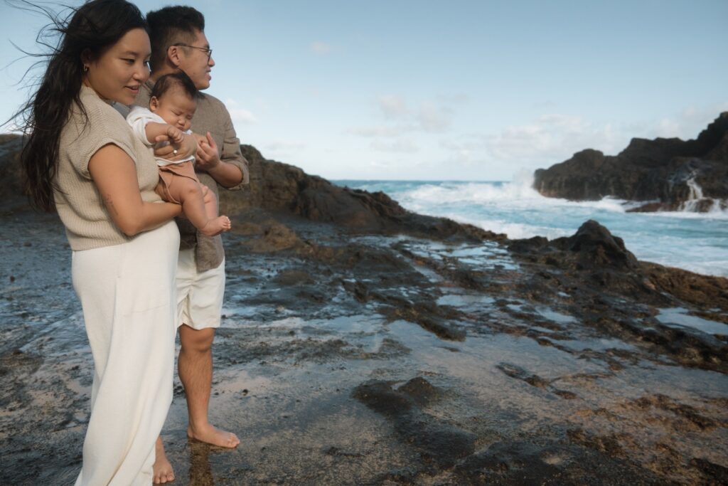 A family photo of a male and female standing at beach cove with baby near Oahu, Hawaii.