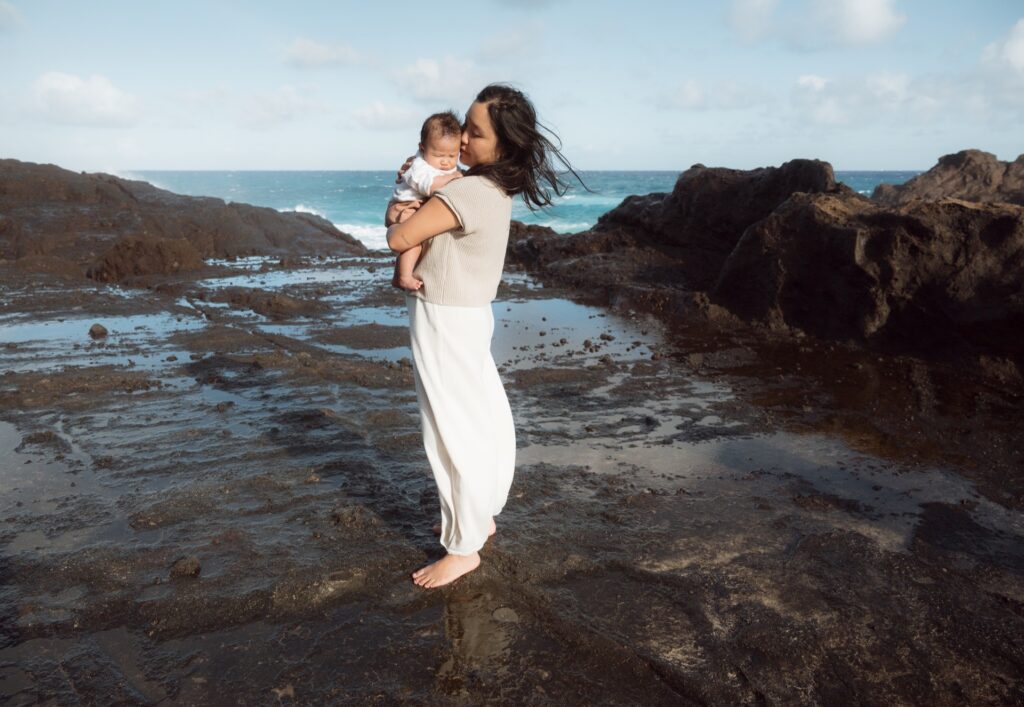 A motherhood portrait of a female standing at beach cove with baby near Oahu, Hawaii.