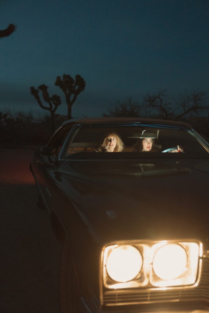 Two females sitting in vintage car with headlights on at night in Joshua Tree National Park.