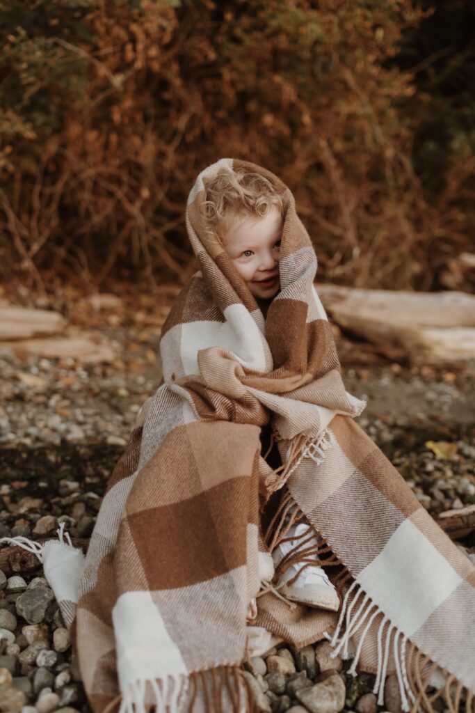 A little boy wrapped in brown checkered blanket sitting on beach near Seattle, WA.