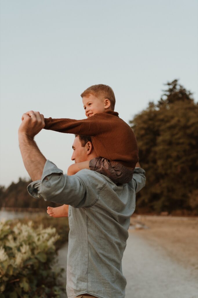 A father holding little boy on shoulders at beach.