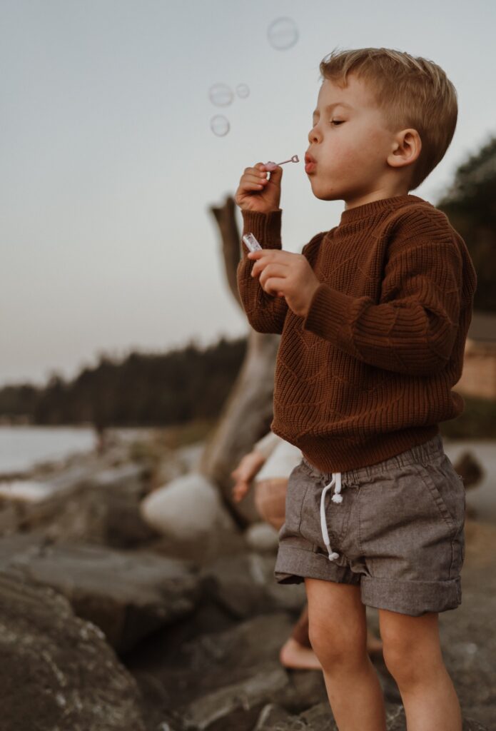 A little boy standing on log at beach blowing bubbles.