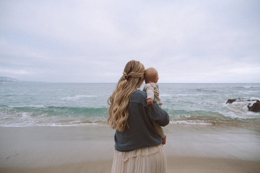 female holding baby wearing cream outfit at laguna beach