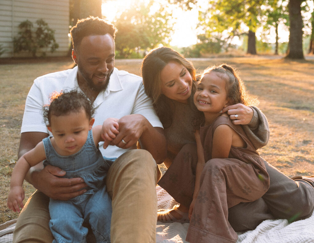 Parents and children sitting on a blanket during golden hour, candid and playful in coordinated neutral outfits.
