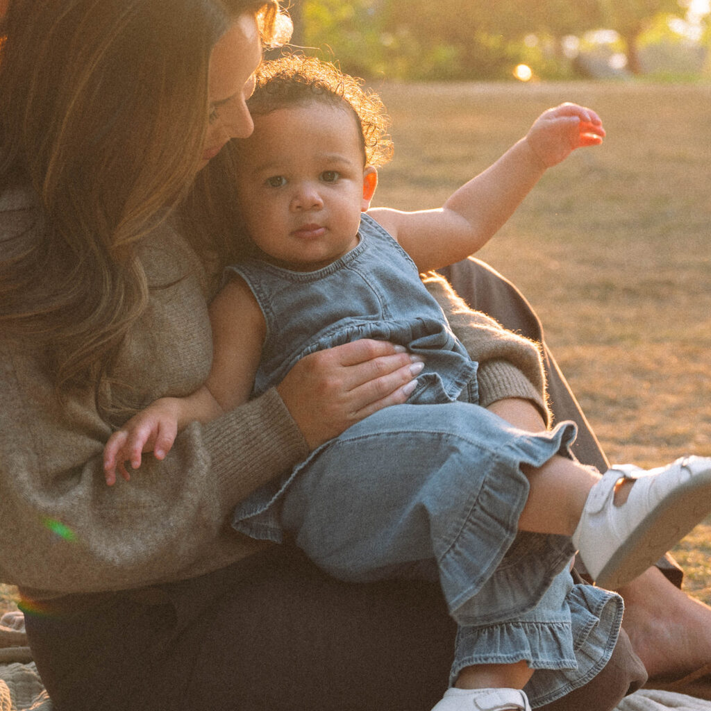 Mother holding toddler in denim jumpsuit during golden hour family session, with warm natural light on their faces.