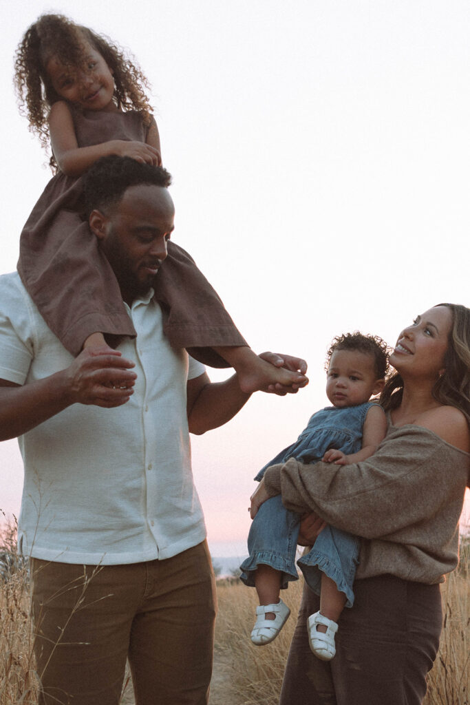 Father carrying daughter on his shoulders while mother holds toddler, styled in earthy neutrals for sunset photos.