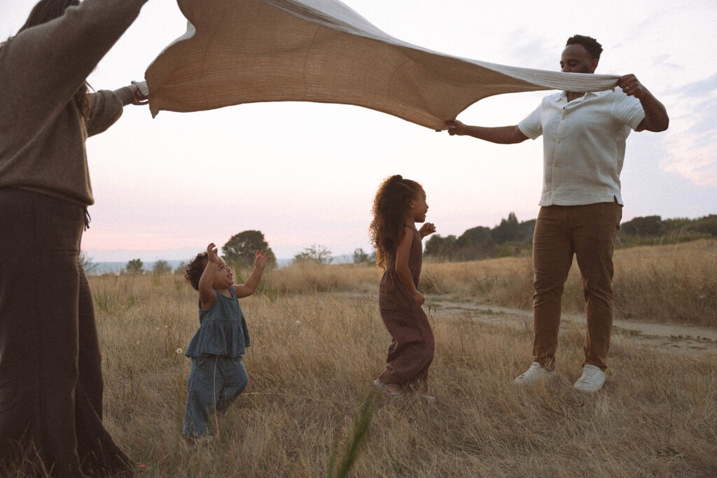 Children playing in a grassy field at golden hour while parents hold up a blanket, capturing candid family photos with natural light.