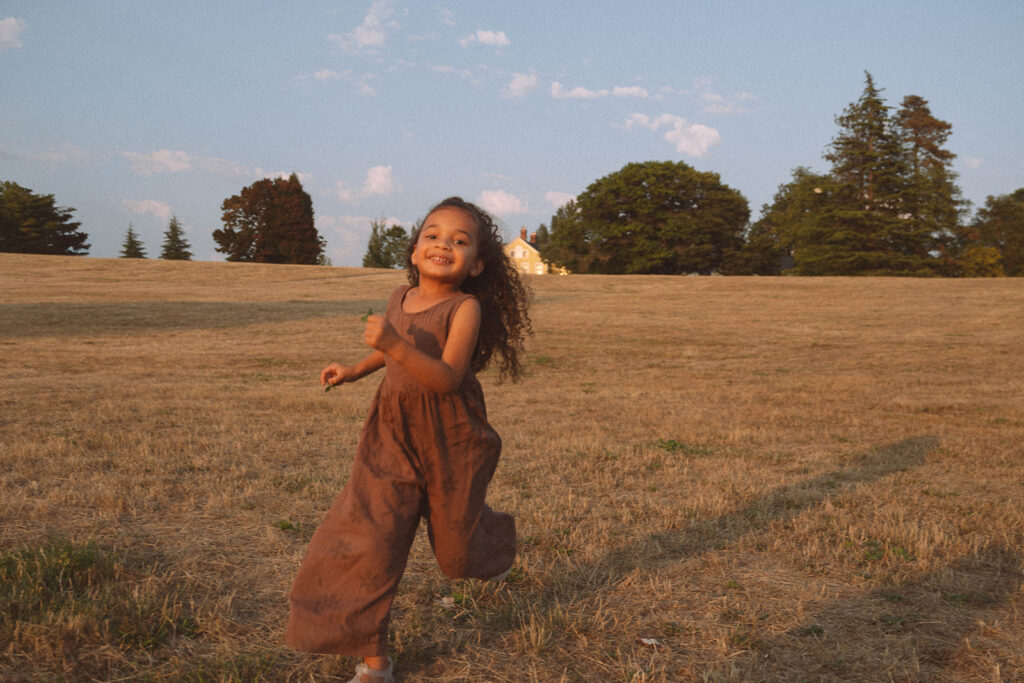 Young girl in a brown jumper running through a golden field at sunset, styled in earthy tones for golden hour family photos.