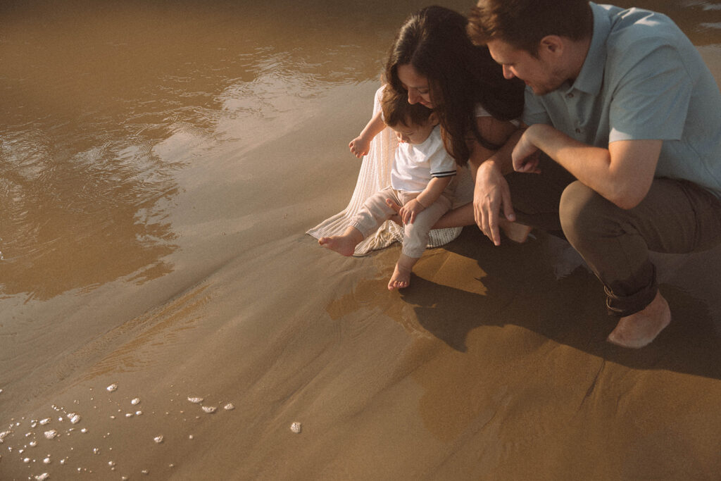 Parents crouching with their baby at the edge of the water during a sunset family session.