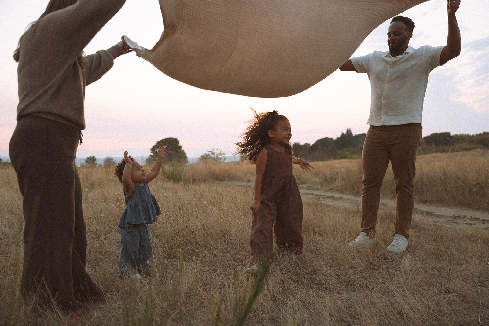 Family playing under a blanket in a golden field at sunset, showing playful candid moments during golden hour.