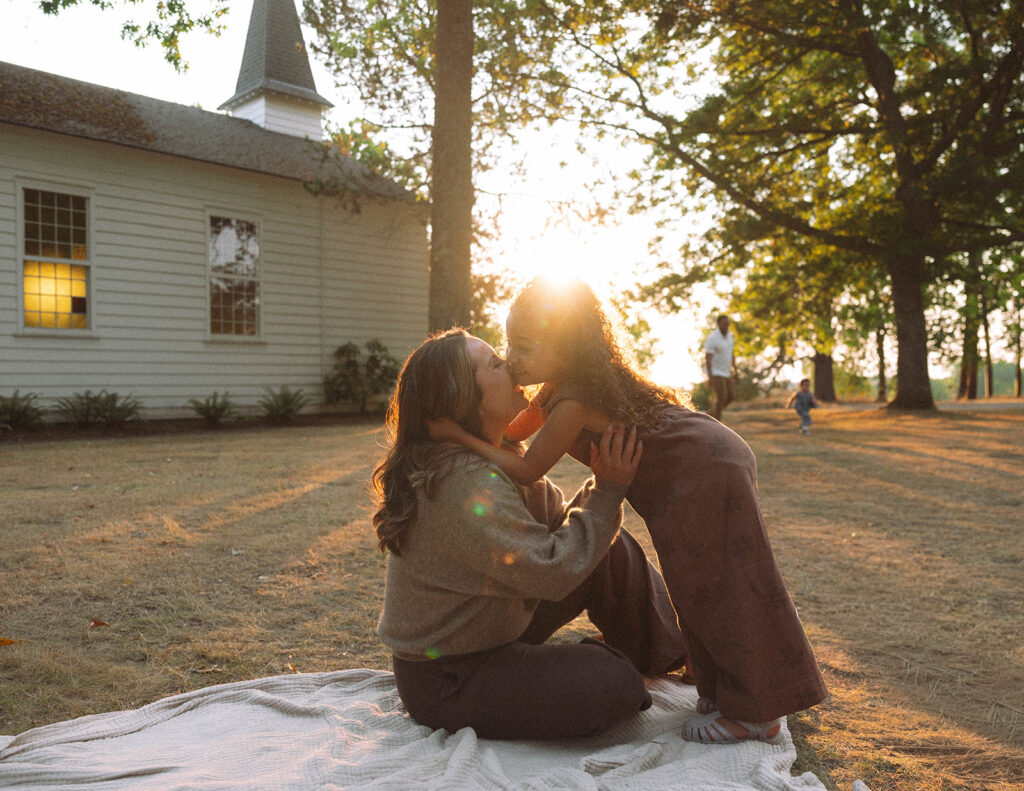 Mother and daughter sharing a kiss on a blanket during golden hour, styled in cozy neutrals and warm light.