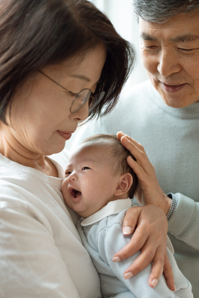 Grandmother cuddling newborn baby while grandfather smiles beside her during an Orange County at-home newborn family session.