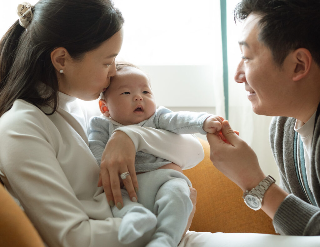 Mother kissing newborn baby while father holds the baby’s hand during a lifestyle newborn session in Irvine, California.