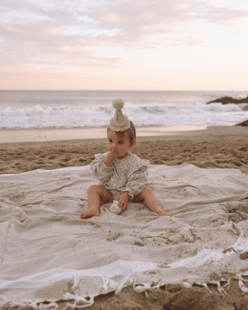 Baby sitting on a beach blanket wearing a tiny birthday hat during a family photoshoot in Laguna Beach, Orange County.