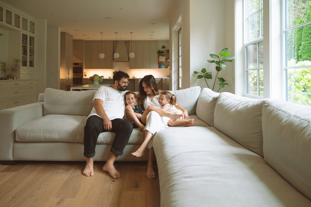 A family sitting together on a large sectional sofa in a bright living room, kids curled up between their parents in a cozy moment.