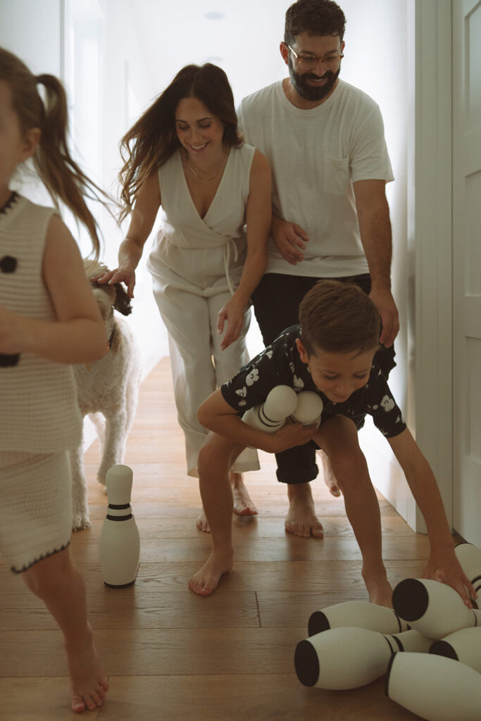 A family playing hallway bowling at home, kids running with pins in hand while parents laugh and join in.