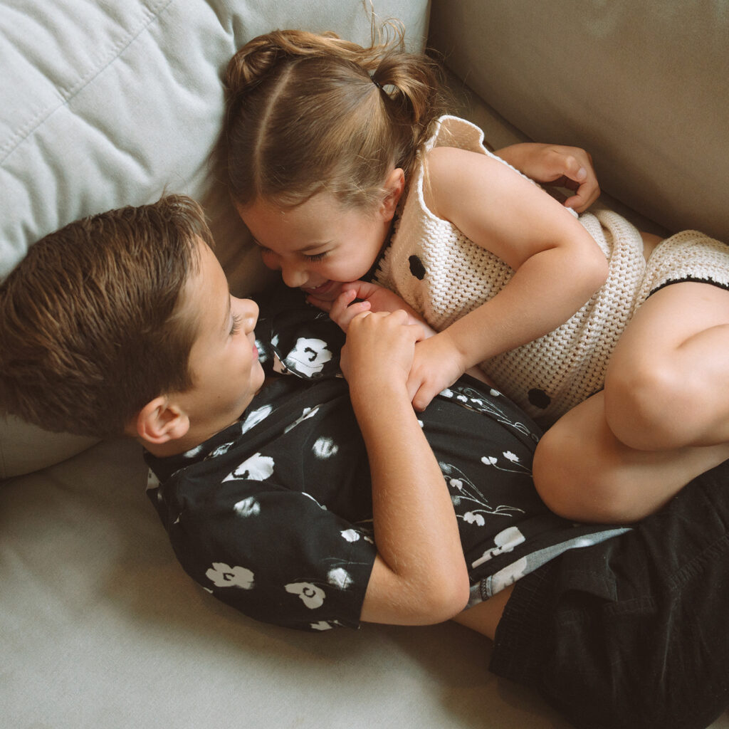 Two siblings lying on a couch, laughing and snuggling together during an at-home family session.