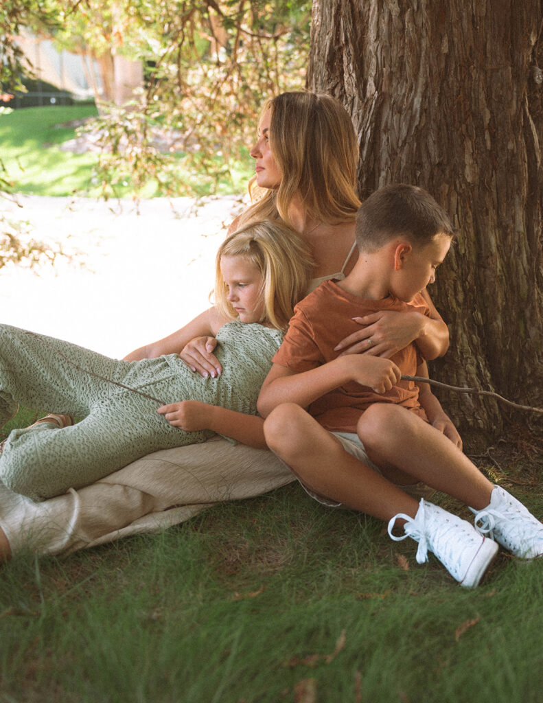 A mom sitting under a large tree with her children nestled closely against her, warm afternoon light filtering through the leaves.
