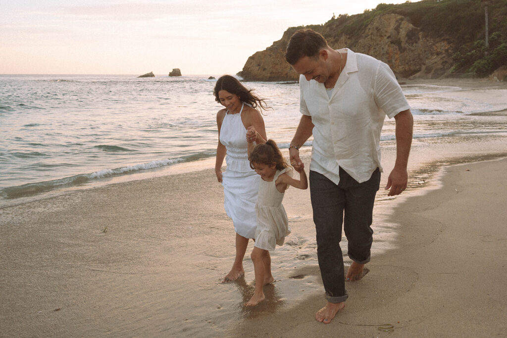 A family walking barefoot along Crescent Bay at sunset, their daughter splashing between them — a warm and candid example of family photos in Orange County by the beach.