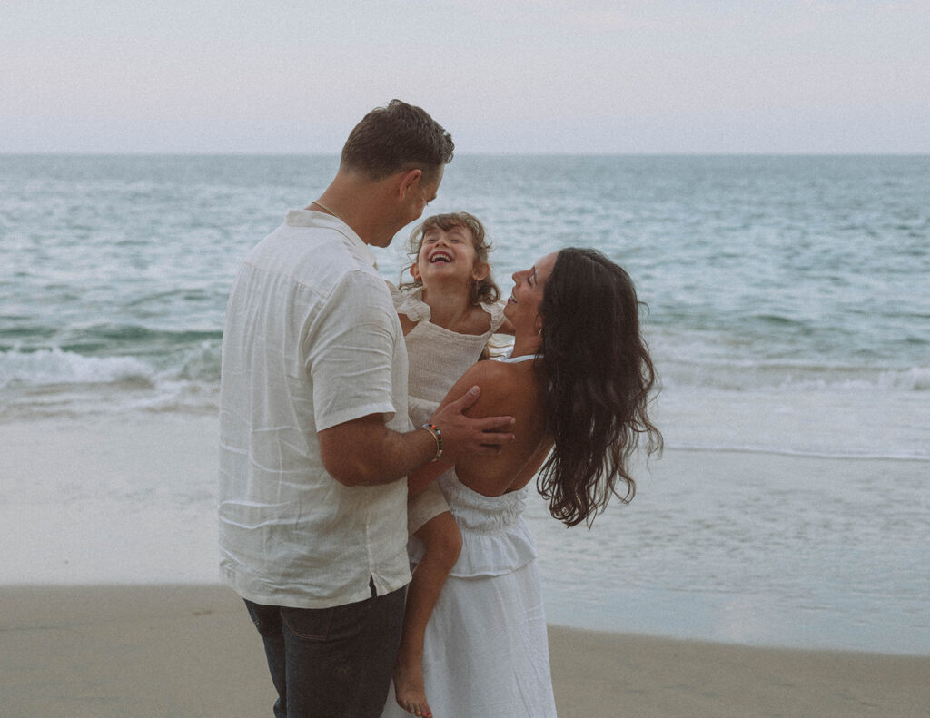Parents holding their daughter at the water’s edge at Crescent Bay, all laughing together against the backdrop of the ocean.