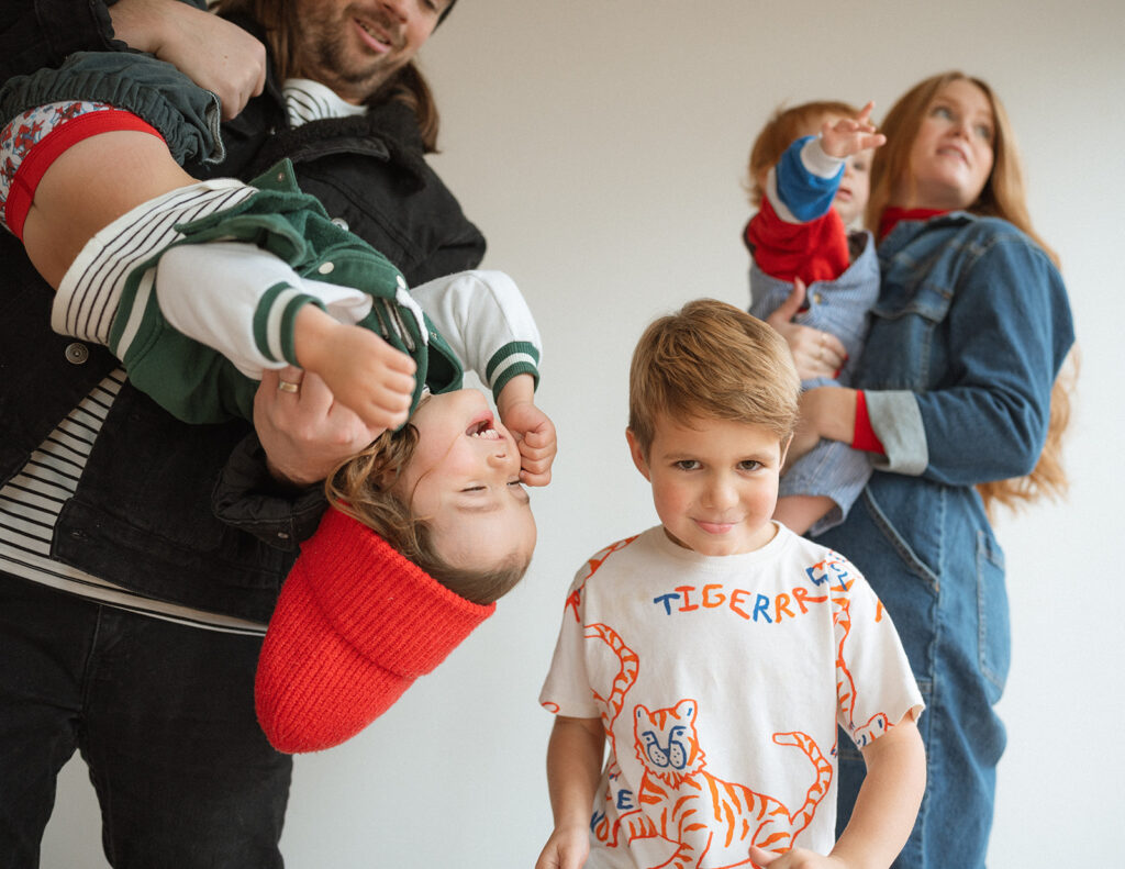 A lively family moment in a studio setting, kids laughing and moving while parents hold younger siblings in the background.