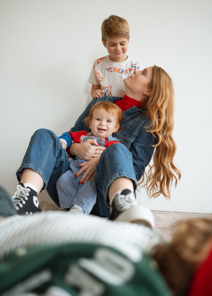 A mom sitting on the floor with her children in a bright studio space, one child laughing in her lap while another looks down at her with a soft smile.