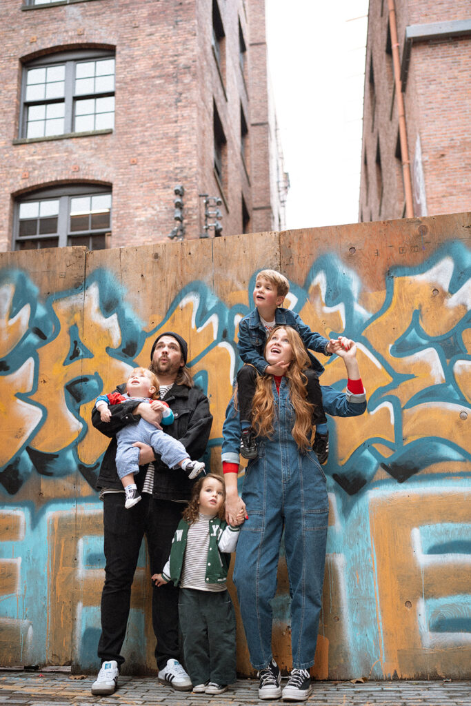 A playful family moment in an urban alley, parents holding their kids in front of a colorful graffiti wall — an example of candid family photos in Orange County with personality and movement.