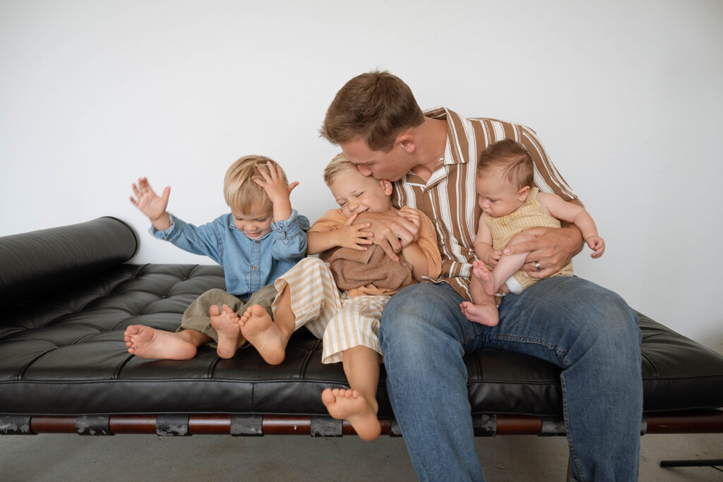 Father sitting with three young children during a relaxed studio family session, captured by photographer Maria Alcantara.