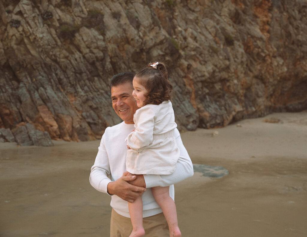 A grandfather holds his young granddaughter while they smile together on the sand at Crescent Bay in Laguna Beach during a South Orange County family session.