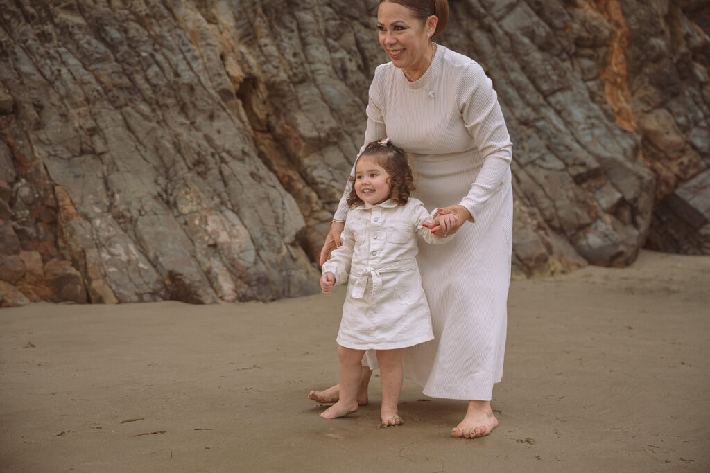 A toddler walks barefoot in the sand at Crescent Bay in Laguna Beach while her grandmother gently guides her during a South Orange County family session.