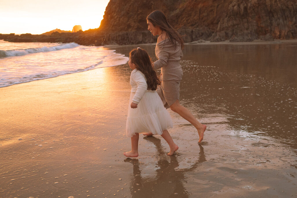 A mother and her young daughter run along the shoreline at Crescent Bay Beach as the sunset reflects warm light across the water in Laguna Beach.