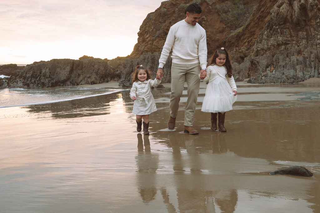 A father walks with his two young daughters along the reflective shoreline at Crescent Bay Beach in Laguna Beach.