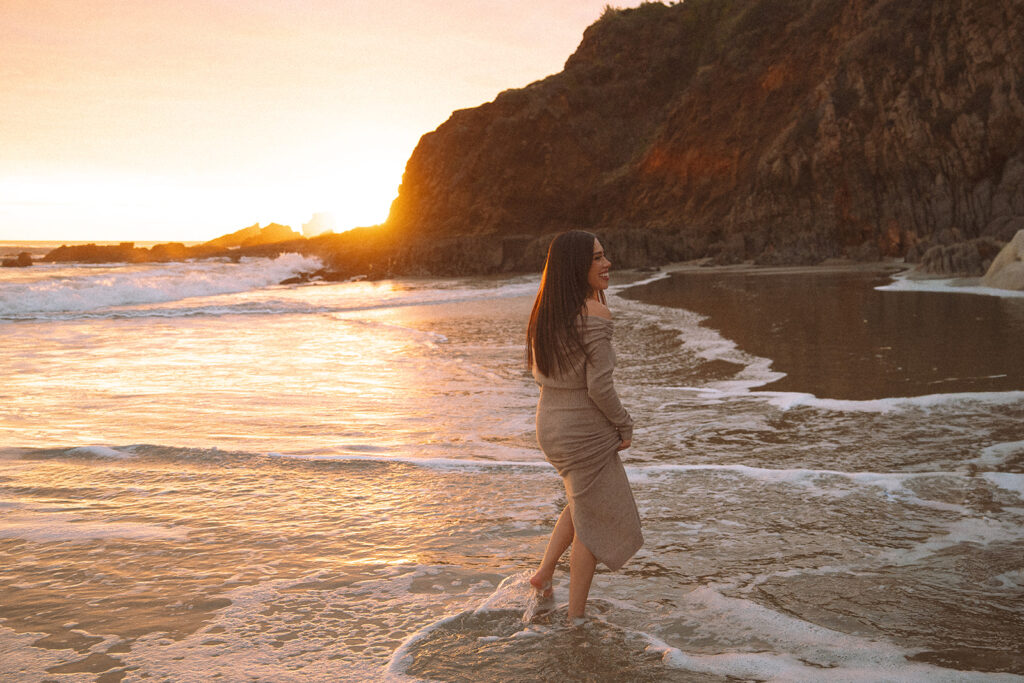 A woman wades through glowing sunset waves at Crescent Bay, surrounded by the rocky coastline of Laguna Beach.