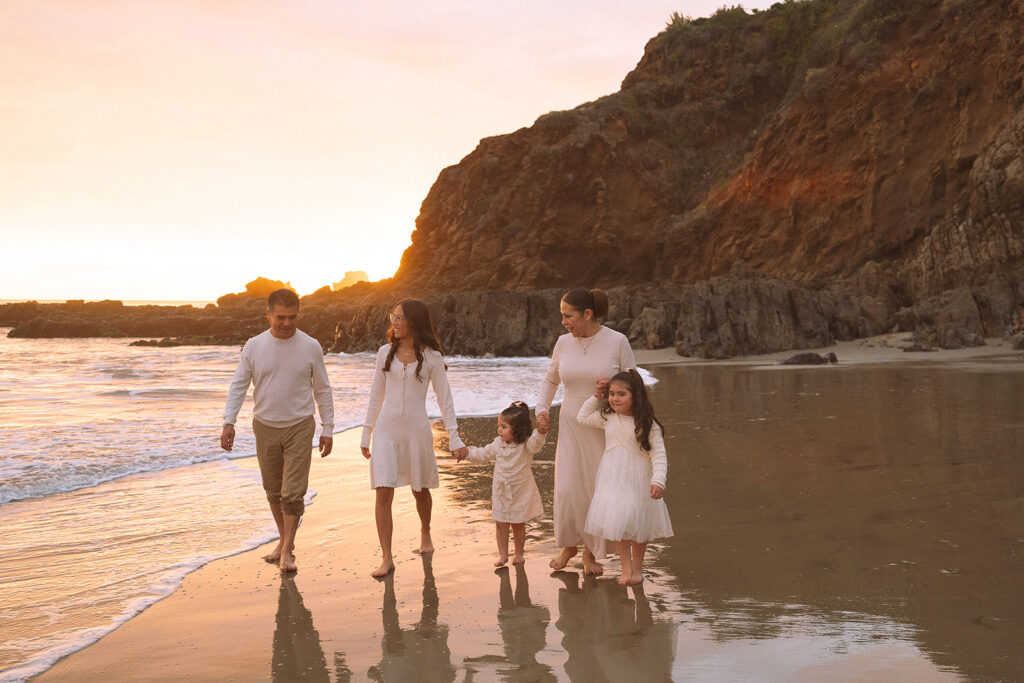 A family walks together along Crescent Bay Beach during golden hour, with soft waves and Laguna Beach cliffs in the background.