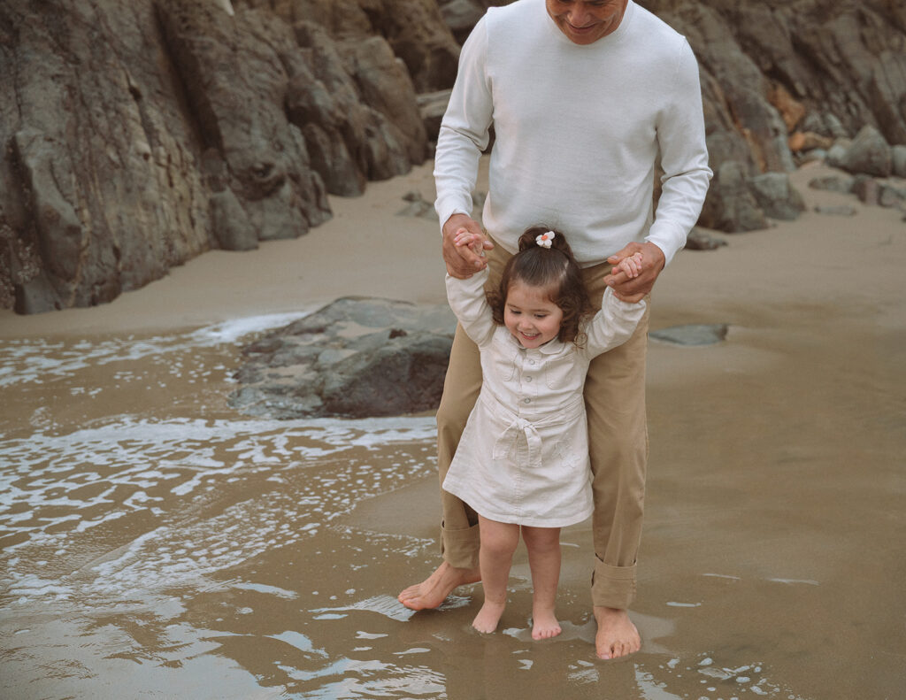 A little girl splashes at the shoreline while her grandfather holds her hands at Crescent Bay Beach in Laguna Beach, captured during a playful South OC family photoshoot.