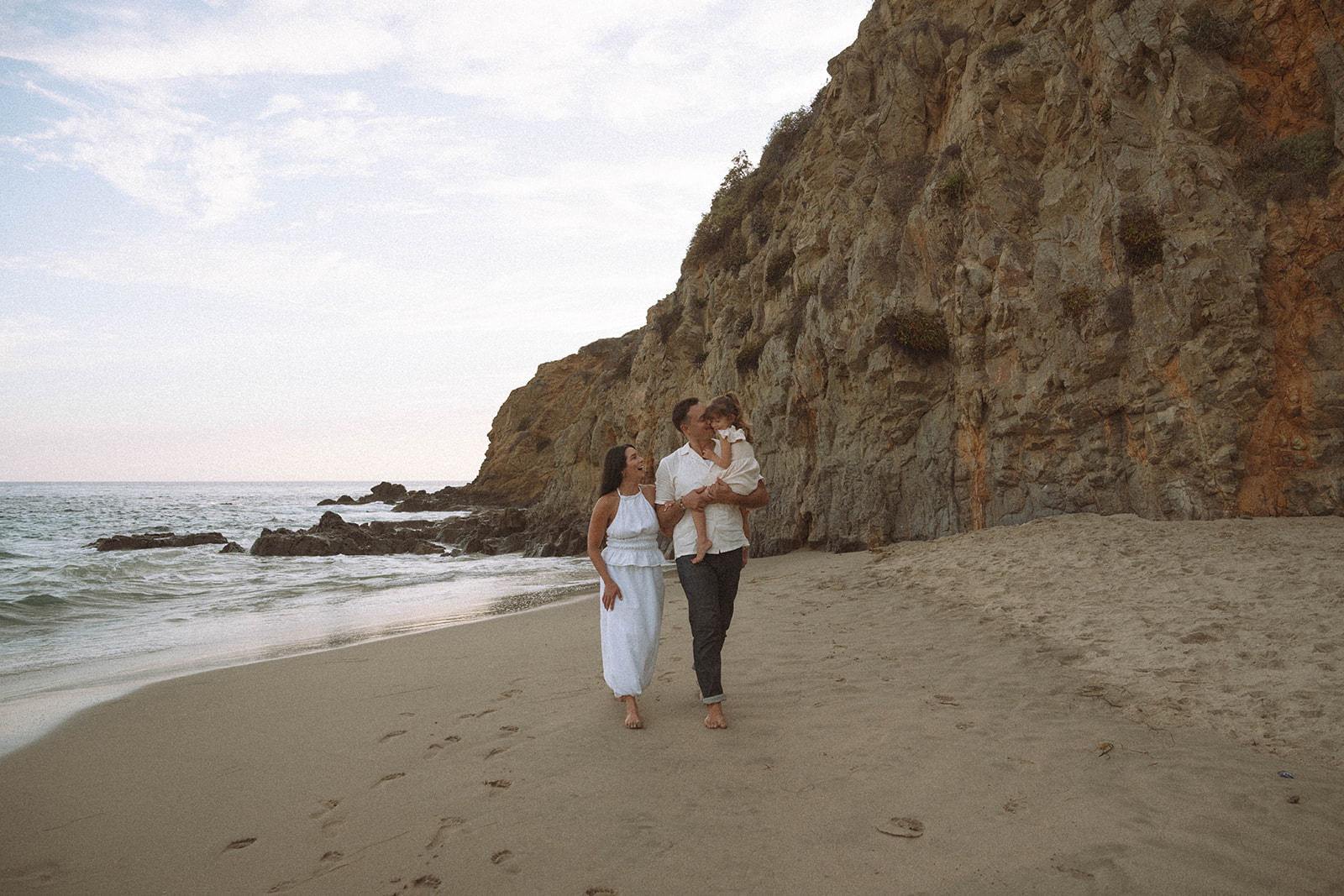 A family walking barefoot along the beach near coastal cliffs during golden hour, photographed during what many families consider the best season for Orange County photos.