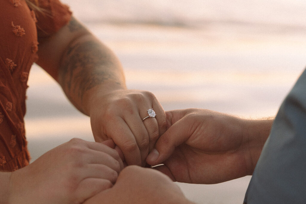 Close-up of an engagement ring during a surprise proposal, with the couple holding hands near the ocean at sunset.