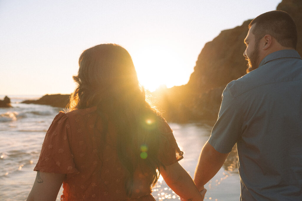 Couple walking hand in hand at during golden hour, backlit by the setting sun and ocean waves.