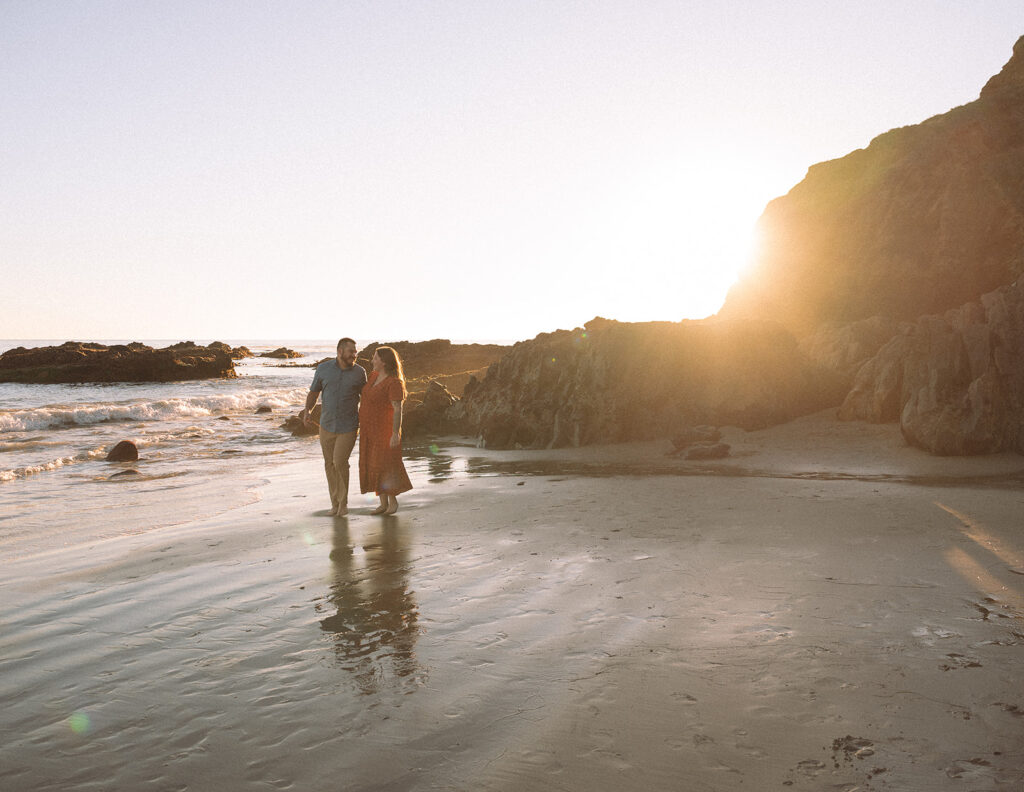 Couple walking along Crescent Bay Beach in Laguna Beach during sunset, with ocean waves, cliffs, and warm golden light in the background.