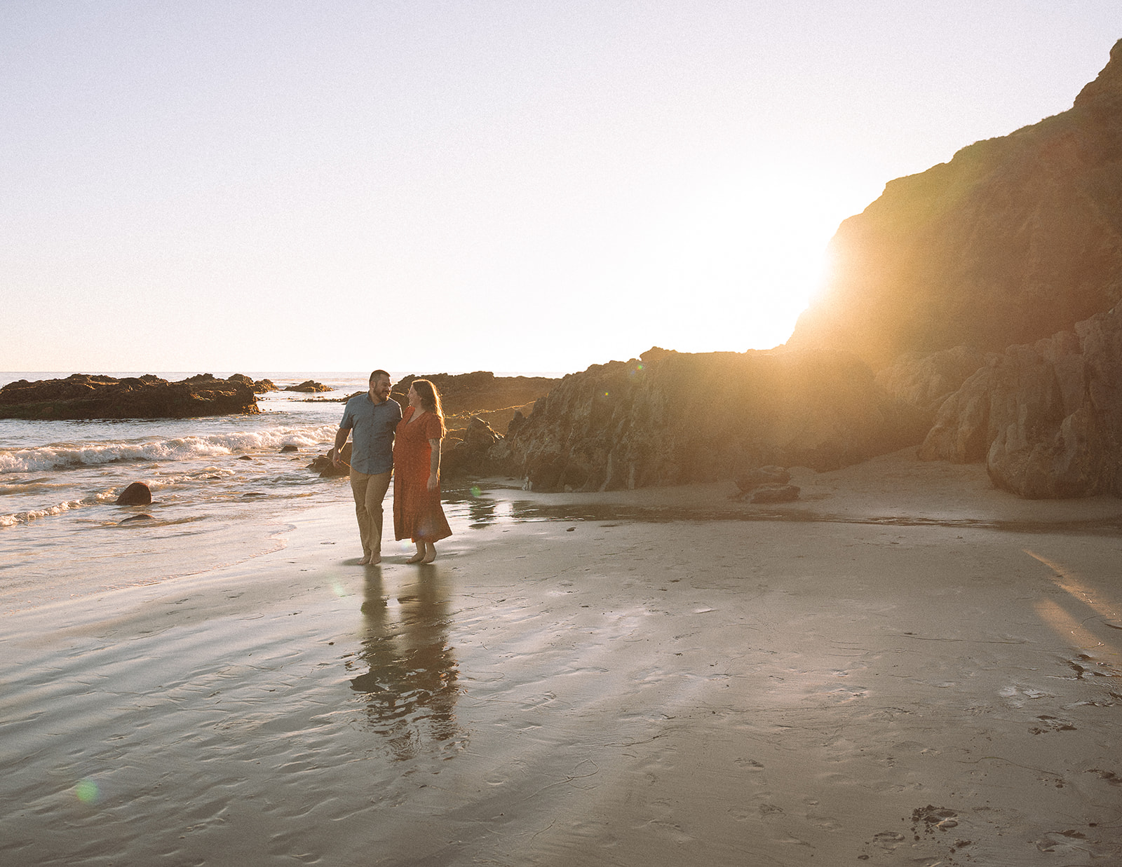 Couple walking along Crescent Bay Beach in Laguna Beach during sunset, with ocean waves, cliffs, and warm golden light in the background.
