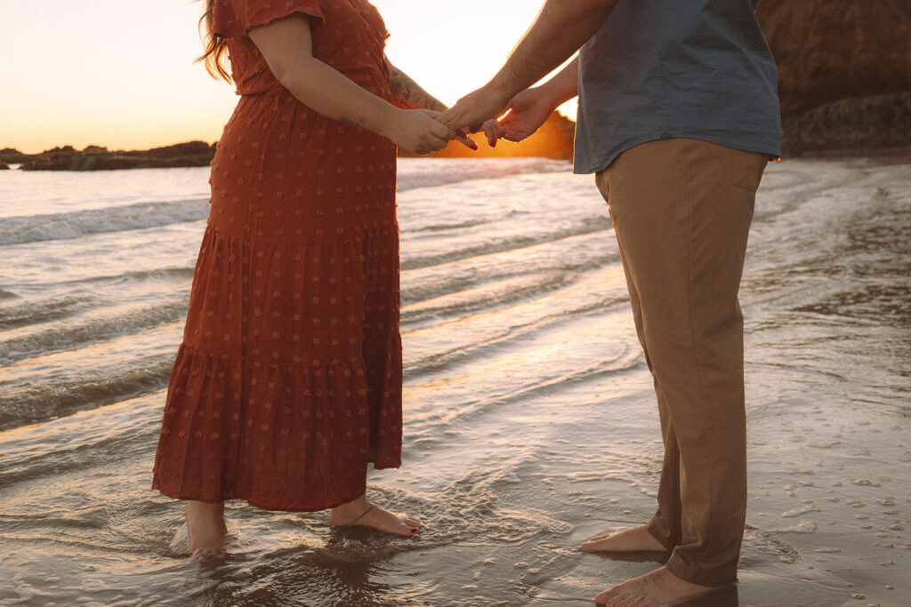 Engaged couple holding hands while walking at a beach during golden hour by the ocean.