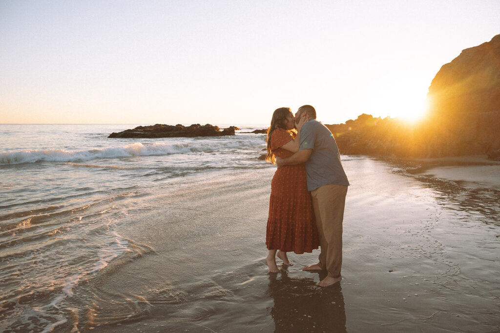 Couple kissing after their surprise engagement standing barefoot at the shoreline during golden hour.