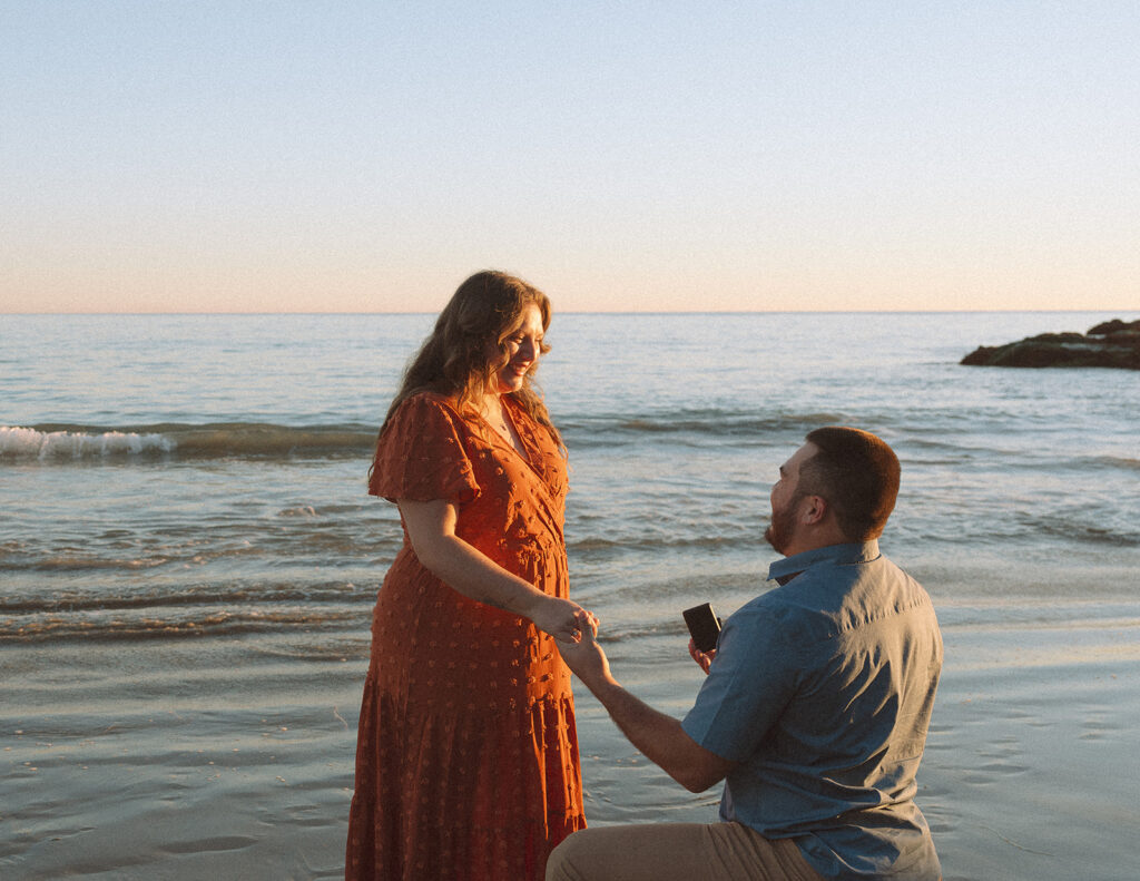 Surprise engagement proposal in Laguna Beach, with one partner kneeling on the shoreline as the other reacts.