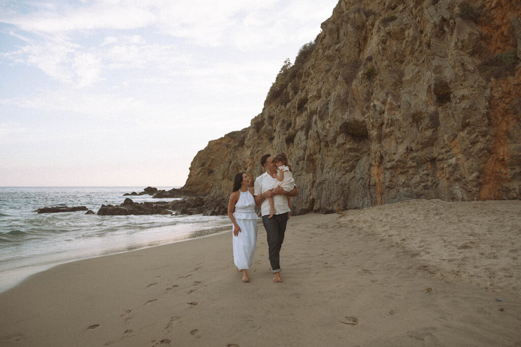 A family walking barefoot along the beach near rocky cliffs, relaxed and unposed.