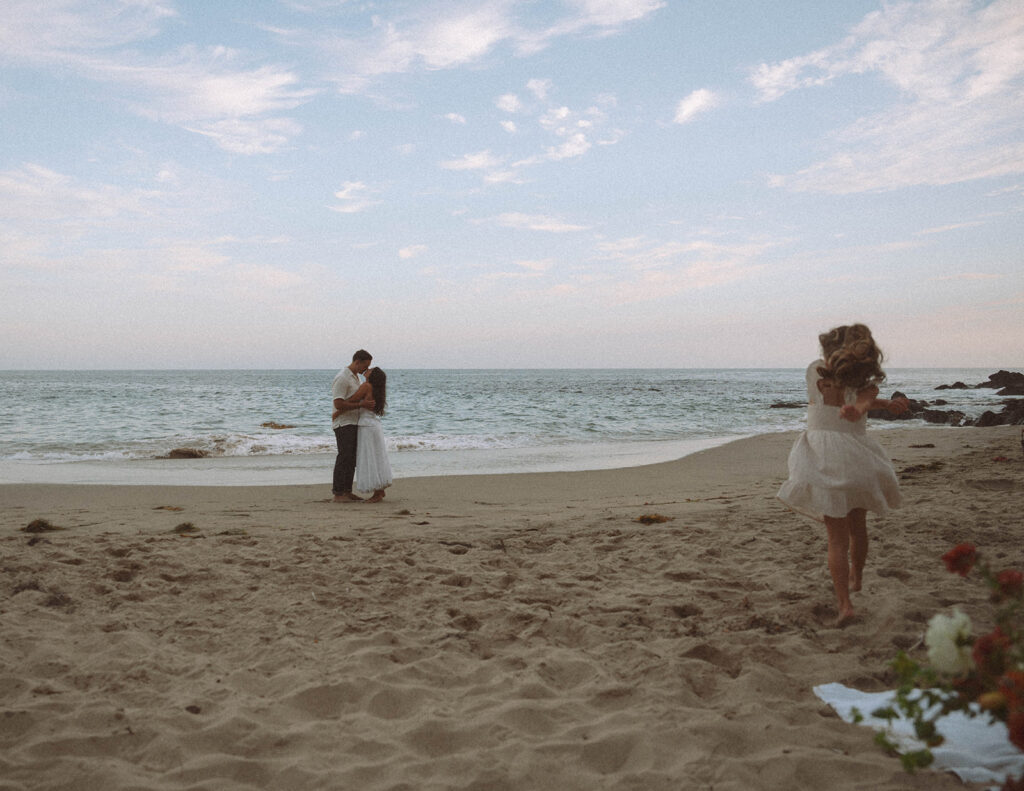 Parents sharing a quiet moment near the shoreline while their child runs toward them across the sand.