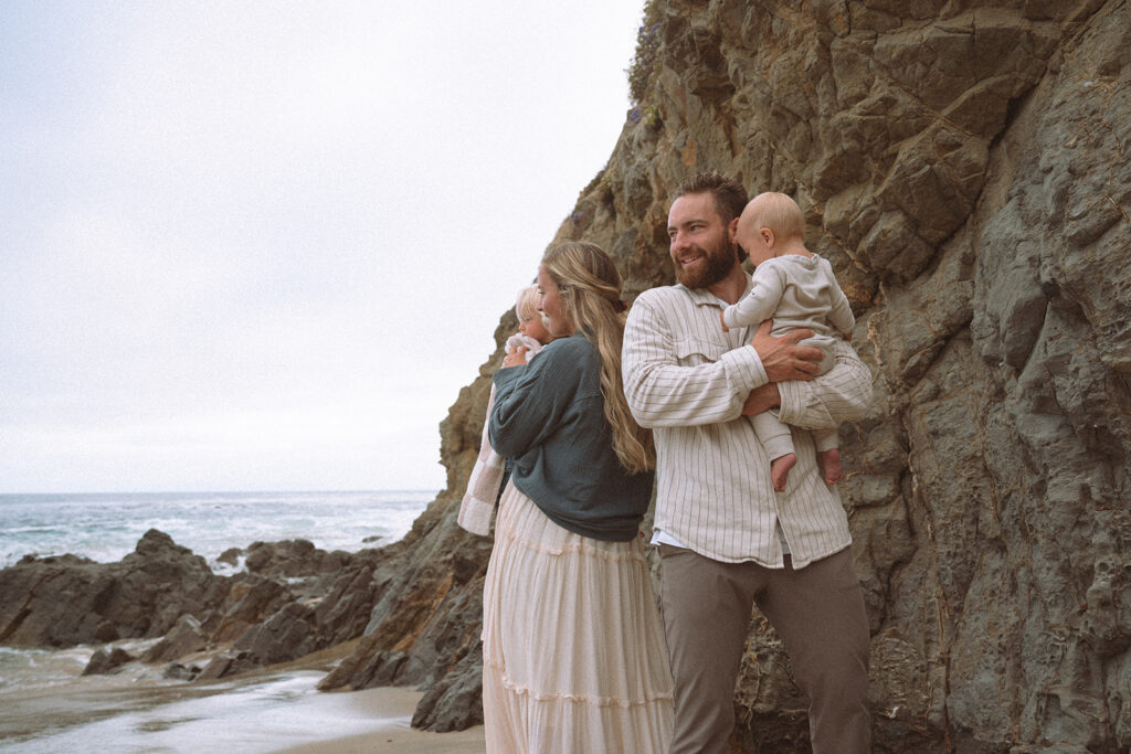 A family standing near the cliffs at the beach, holding their baby and looking out toward the ocean on a quiet day.